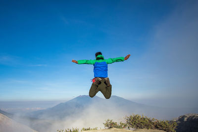 Rear view of man jumping on mountain against blue sky