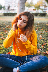 Young girl in yellow sweater listening to the music with earphones sitting on autumn grass
