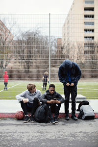 Phone addicted friends on sidewalk against playing field in city