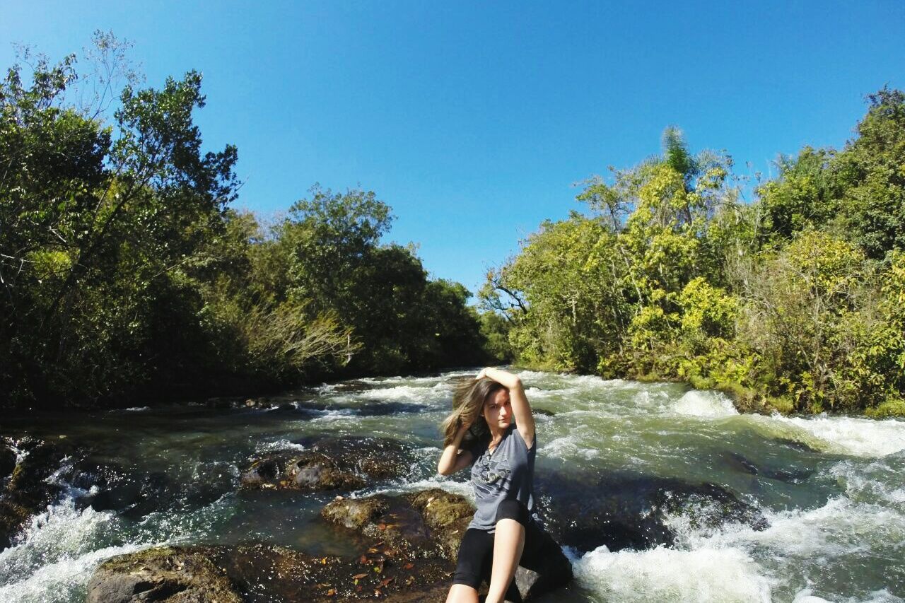 tree, real people, water, one person, leisure activity, young adult, nature, day, young women, rock - object, beauty in nature, river, sitting, forest, outdoors, clear sky, lifestyles, waterfall, sky, people