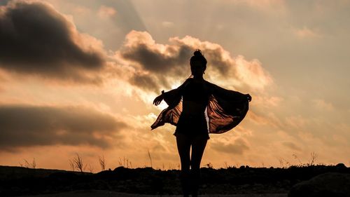 Silhouette woman with arms outstretched standing on land against sky during sunset