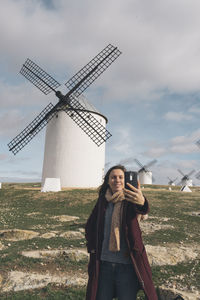 Woman taking selfie while standing against traditional windmills on landscape