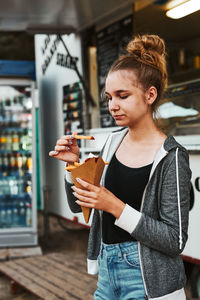 Teenage girl eating potato fries from carton cone standing in front of food truck