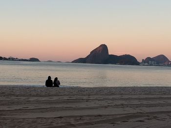 Silhouette people sitting on beach against clear sky during sunset