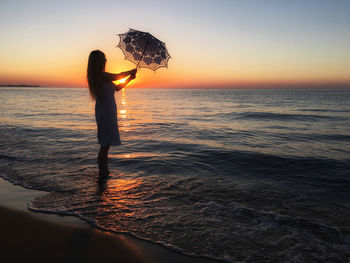 Side view of silhouette woman with umbrella standing at beach against sky during sunset