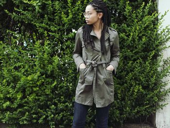 Young woman with hands in pockets standing against plants