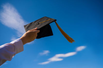 Low angle view of woman holding mortarboard against blue sky