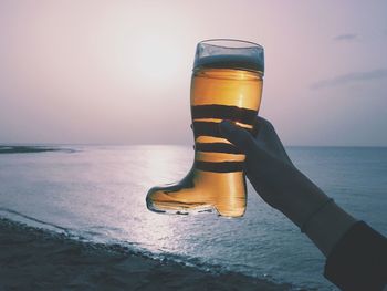 Cropped hand of woman holding boot shape beer glass at beach against sky during sunset