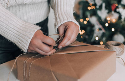 Midsection of woman wrapping christmas present in kraft paper in front of christmas tree at home
