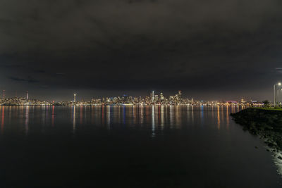 Illuminated city by sea against sky at night