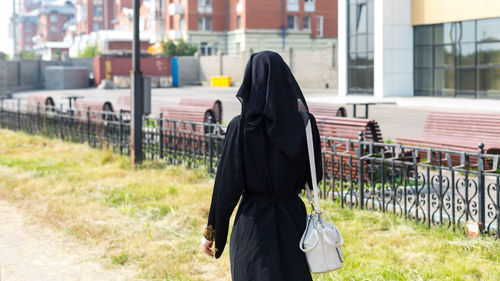A muslim woman walking down a city street in a black national outfit view from the back.