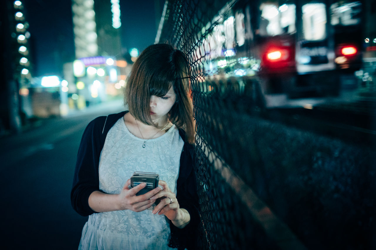 technology, wireless technology, city, smart phone, mobile phone, standing, architecture, one person, illuminated, night, portable information device, using phone, real people, focus on foreground, casual clothing, young adult, lifestyles, street, connection, communication, hair, hairstyle, outdoors, beautiful woman