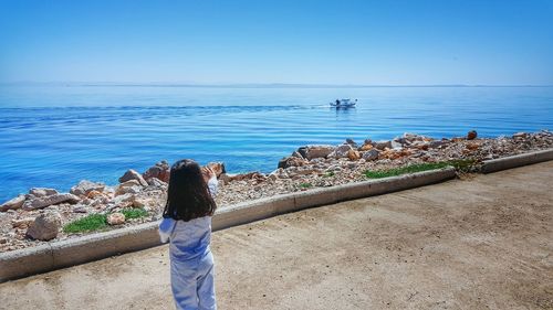 Rear view of little girl waving on promenade by sea against clear sky