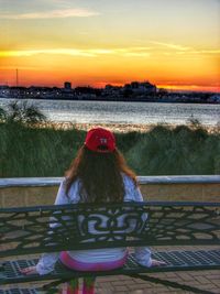 Rear view of woman sitting on railing against sky during sunset