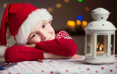 Close-up portrait of cute boy wearing santa hat sitting by oil lamp at home