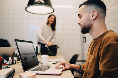 Side view of young male hacker using laptop while female colleague working at small office