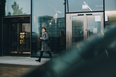 Businesswoman with smart phone looking away while walking on sidewalk against building in city