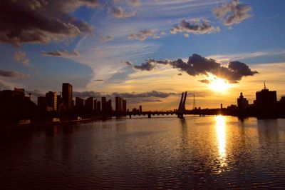 Silhouette buildings by river against sky during sunset