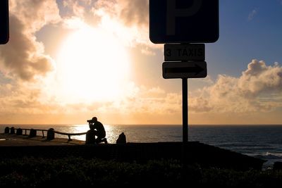 Silhouette photographer photographing at sea by information sign against sky