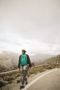 Rear view of young man walking on road while looking at mountains holding skateboard against sky