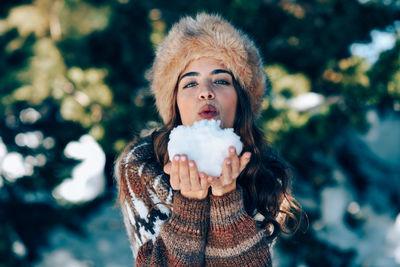 Portrait of young woman holding snow while standing against trees during winter