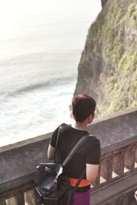 Rear view of man sitting on rock by sea against sky