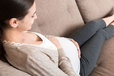 Photo of cute caucasian future mom lying in light living room on sofa and holding hands on tummy.