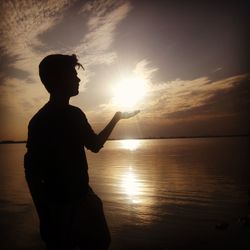 Optical illusion of silhouette man holding sun at lakeshore during sunset
