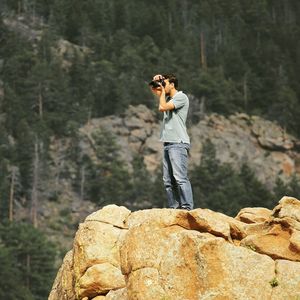 Man photographing with camera while standing on rock against trees in forest