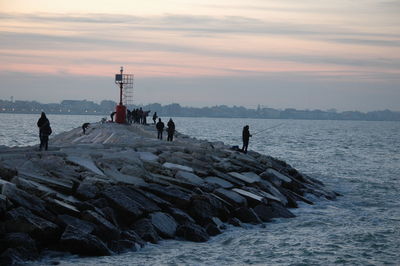 People on rocks by sea against sky during sunset