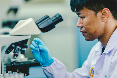 Asian man lab technician in protective glasses and gloves sits next to a microscope in laboratory.
