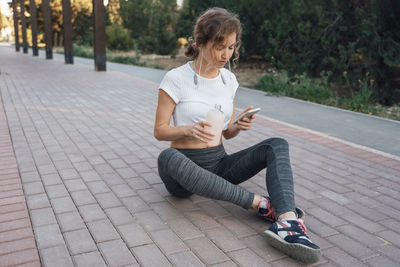 Full length of woman using mobile phone while sitting on footpath