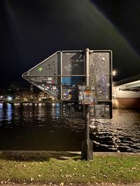 Illuminated sign by river in city against sky at night