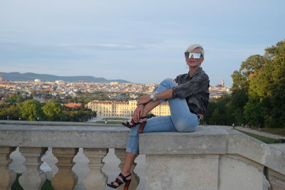 Portrait of woman sitting on stone railing with sunglasses against cityscape