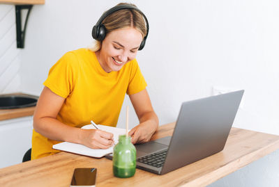 Young smiling blonde woman in yellow t-shirt with headphones working at laptop in kitchen at home
