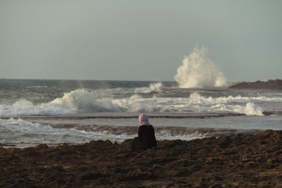 Rear view of woman sitting on rocky shore against clear sky