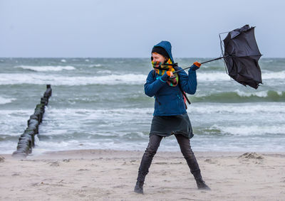 Woman holding damaged umbrella while standing at shore of beach against sky