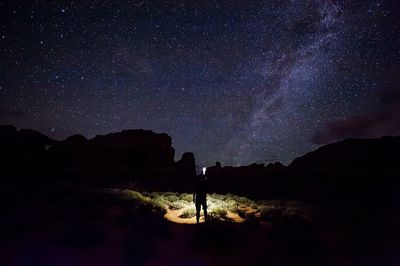 Silhouette hiker with flashlight standing by rocky mountains against star field