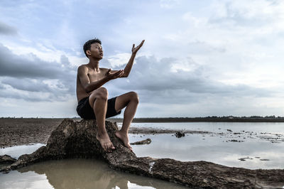 Shirtless boy sitting on rock against cloudy sky during arid climate