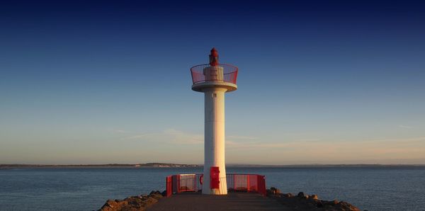 Lighthouse by sea against clear sky during sunset