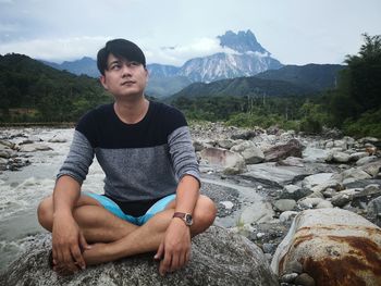 Full length of young man looking away while sitting on rock against sky
