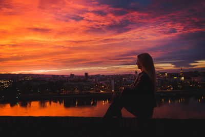 Woman sitting on river with city in background at sunset