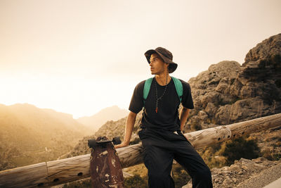 Young male explorer looking away while sitting on railing by skateboard against sky