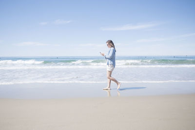 Full length side view of mature woman using mobile phone while walking at beach