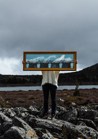 Person holding mirror with reflection while standing on land against cloudy sky