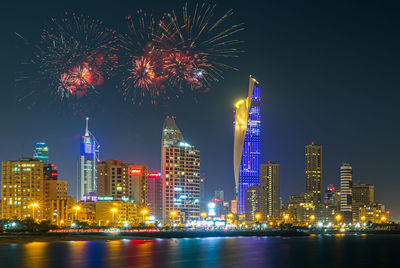 Firework display over river by illuminated buildings against sky at night