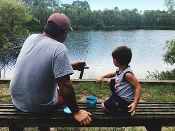 Father and son sitting on bench while fishing at lake