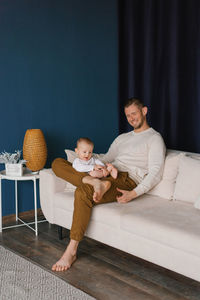 A happy young dad holds his six-month-old son in his arms, sitting on the sofa in the living room