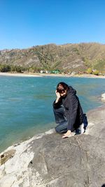 Young woman sitting on rock against blue sky