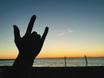 Silhouette hand gesturing horn sign against sea during sunset